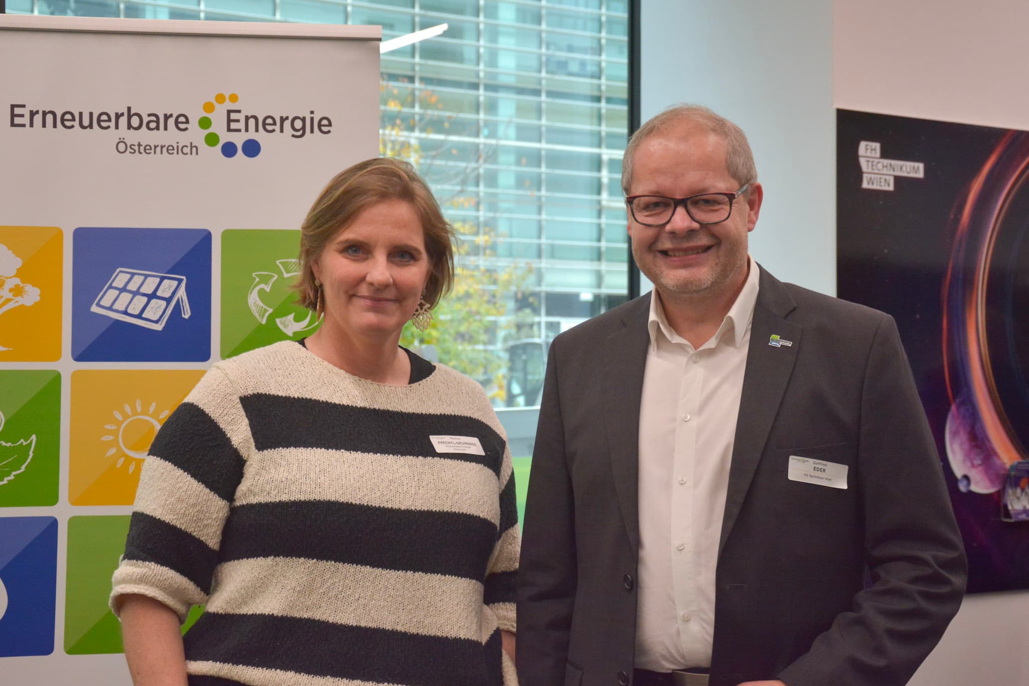 Zwei Personen stehen in einem Haus und lächeln in die Kamera. Hinter ihnen ist ein Banner für Erneuerbare Energie Österreich mit bunten Symbolen und Text zu sehen, und ein Fenster, das ein Bürogebäude im Freien zeigt.
