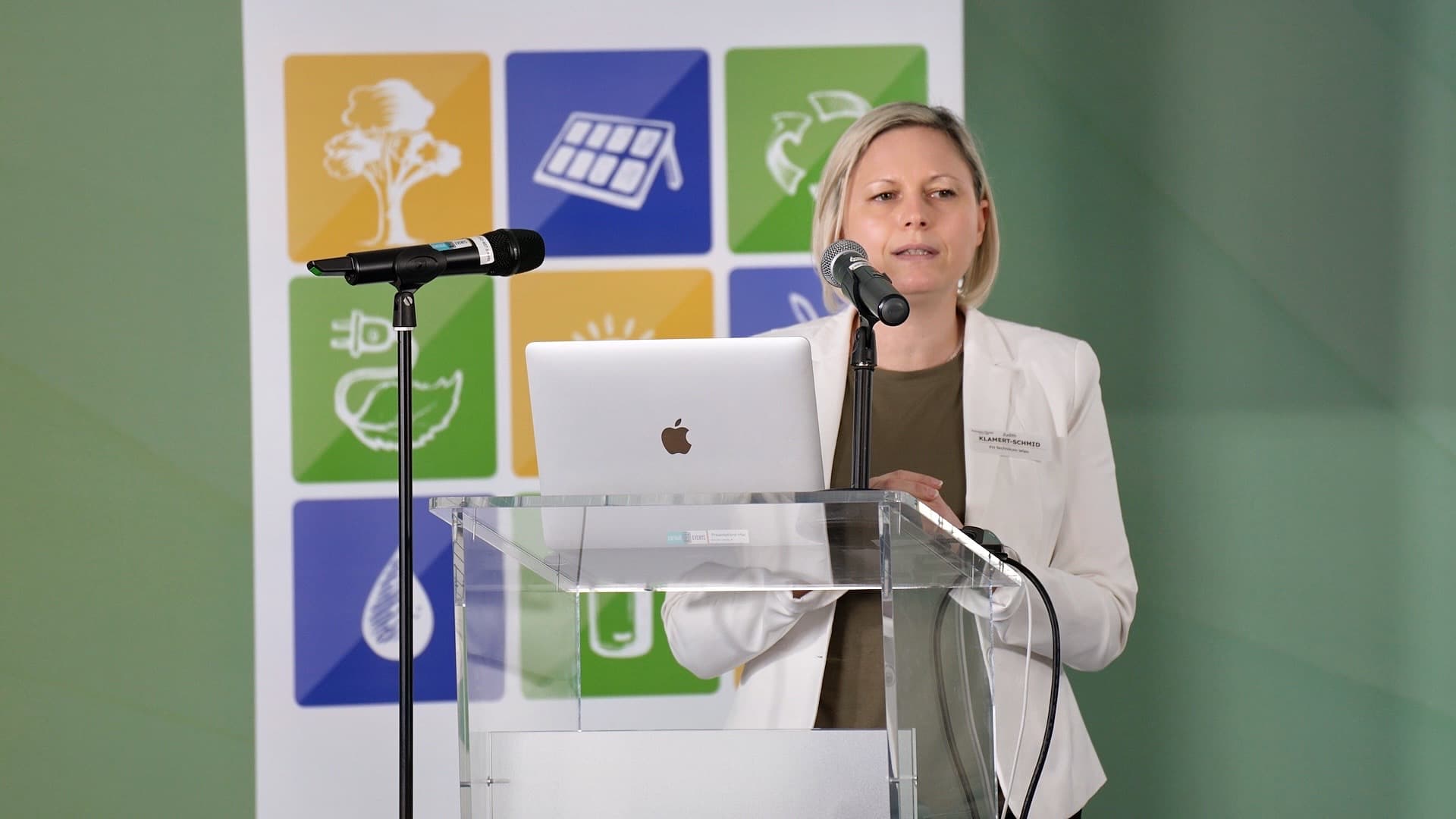 Eine Frau in einem weißen Blazer steht an einem durchsichtigen Podium mit einem Laptop und zwei Mikrofonen und spricht. Hinter ihr hängt ein Banner mit bunten Symbolen, die für erneuerbare Energien und Nachhaltigkeit stehen.