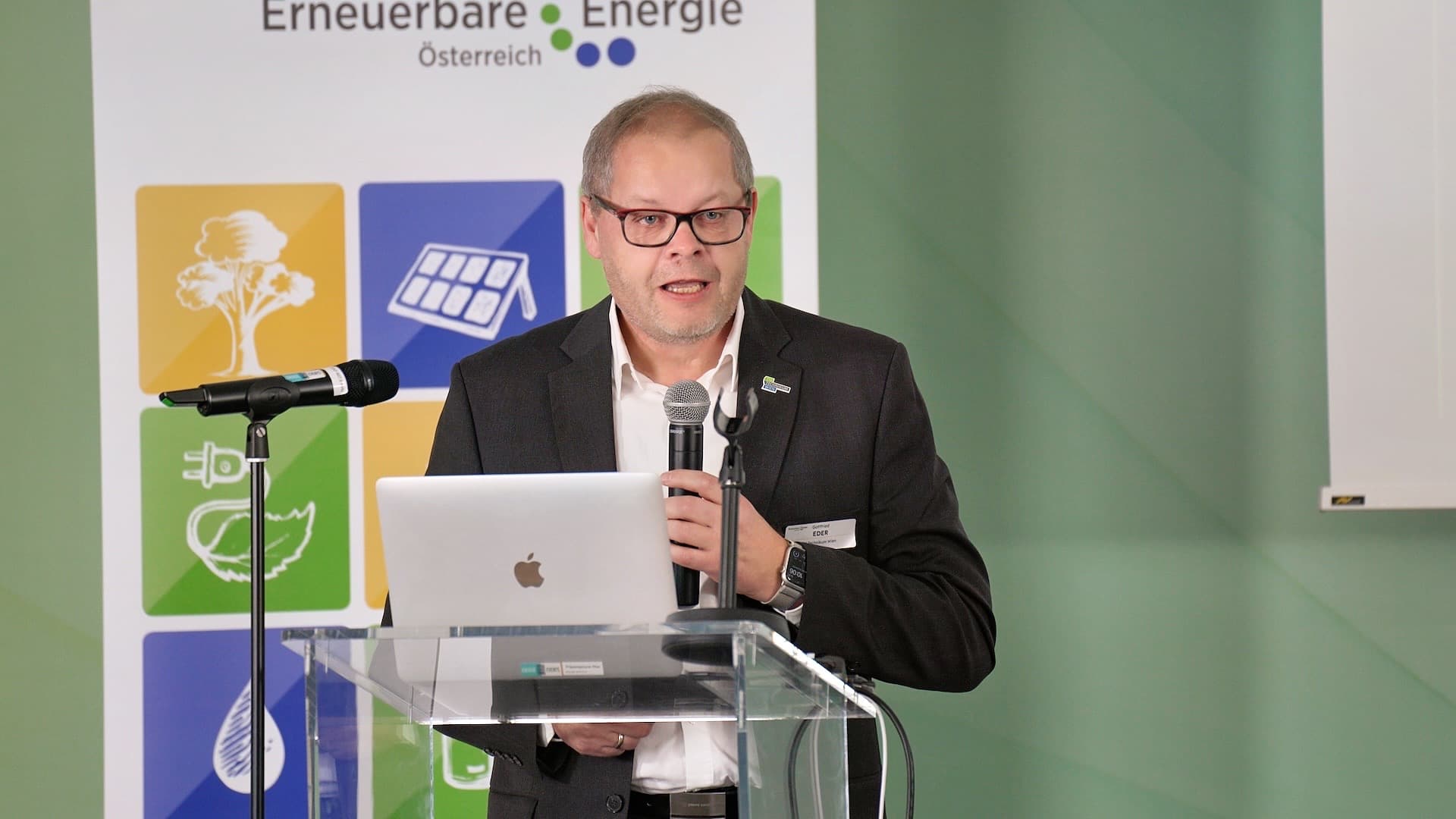 Ein Mann im Anzug spricht auf einem Podium mit einem Laptop in ein Mikrofon. Hinter ihm ist ein Transparent mit Symbolen für erneuerbare Energie und dem Text Erneuerbare Energie Österreich zu sehen.