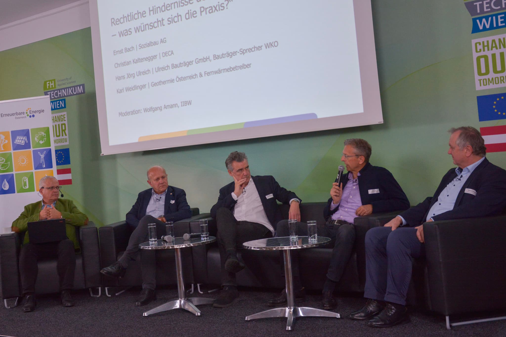 Fünf Männer sitzen auf einer Bühne in einer Podiumsdiskussion, sprechen und gestikulieren. Auf einem Bildschirm hinter ihnen wird ein Präsentationstext in deutscher Sprache angezeigt, und im Hintergrund sind Banner mit Logos und Slogans zu sehen.