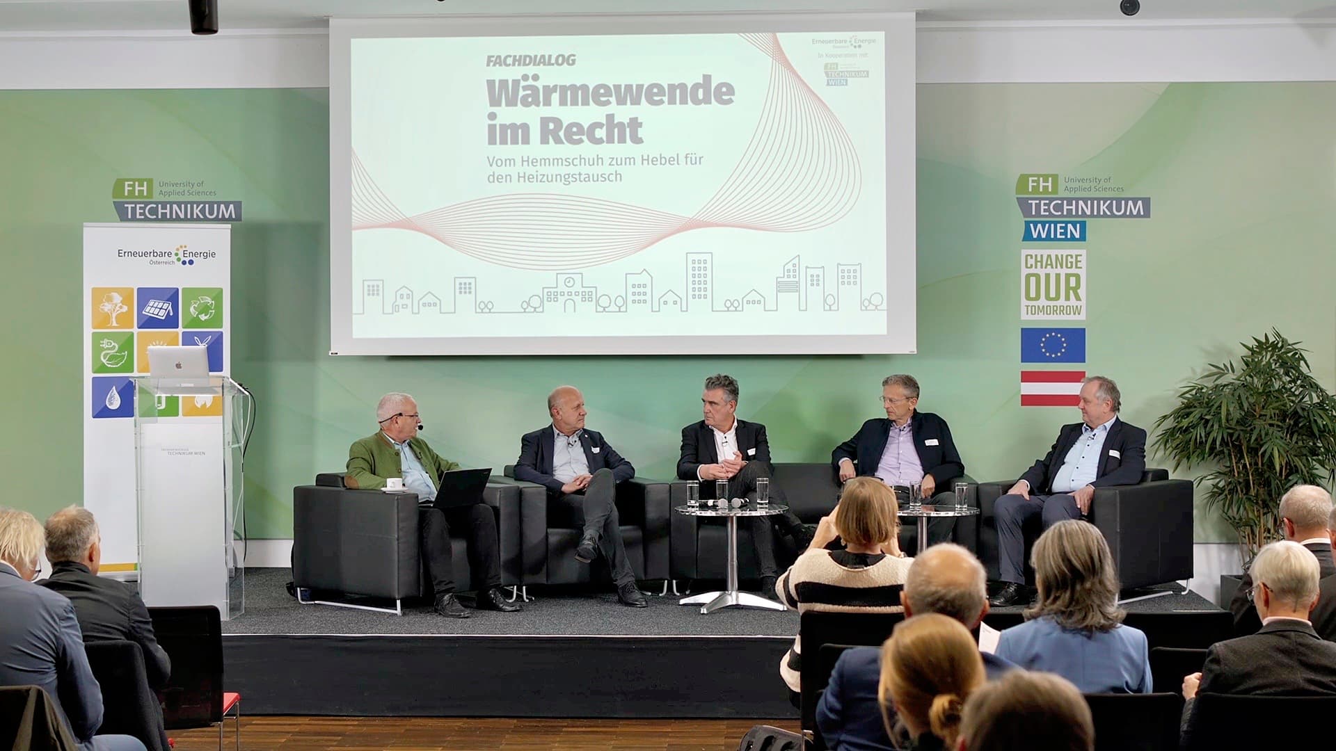 Fünf Personen sitzen auf einer Bühne und führen eine Podiumsdiskussion vor einem Publikum. Eine große Leinwand hinter ihnen zeigt das Thema 'Wärmewende im Recht'. An den Seiten der Bühne sind Plakate mit Symbolen und Fahnen zu sehen.