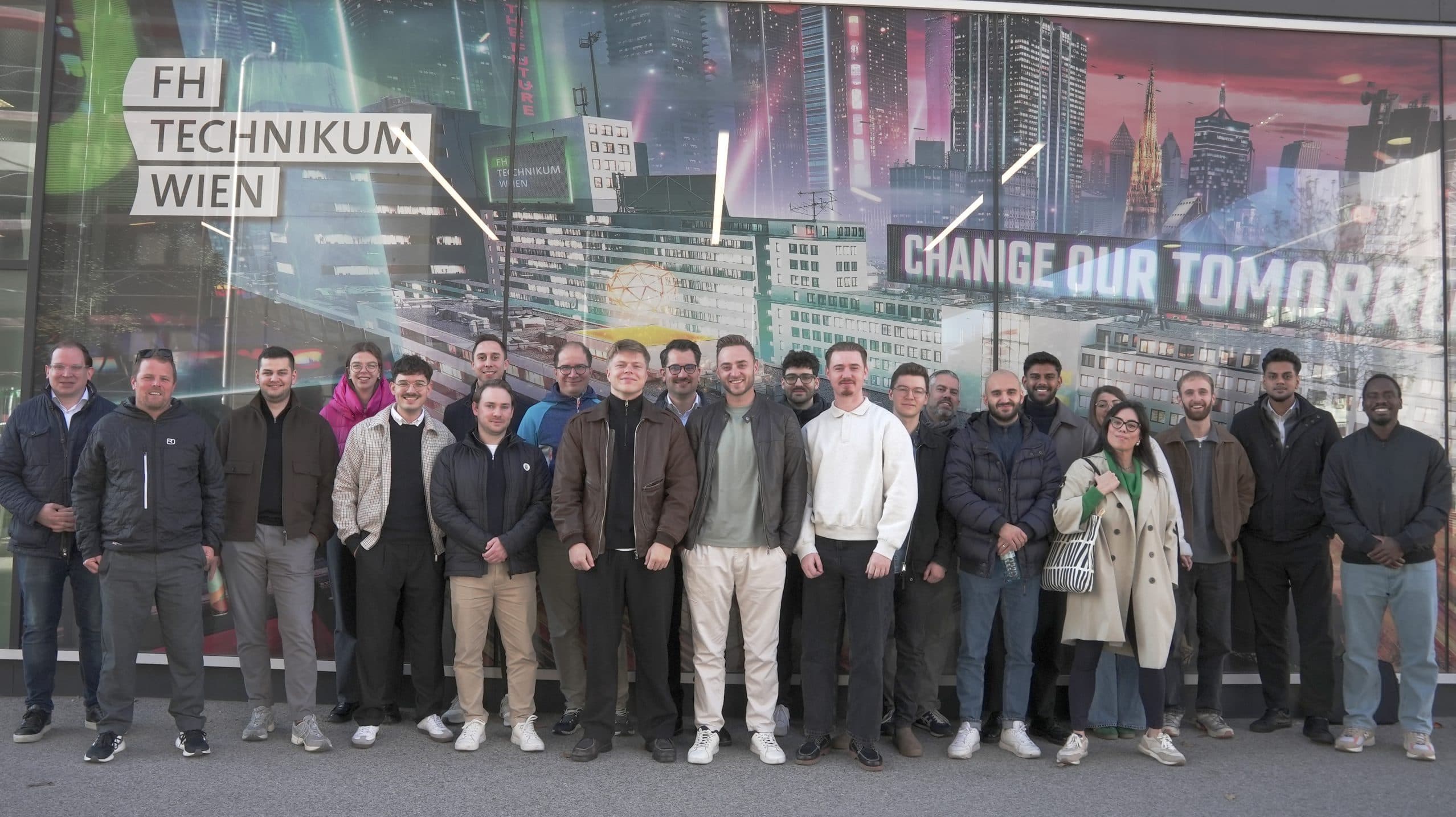 Eine Gruppe von etwa zwanzig Personen steht in einer Reihe und lächelt für ein Foto vor einer futuristischen Stadtlandschaft mit der Aufschrift FH TECHNIKUM WIEN und CHANGE OUR TOMORROW.