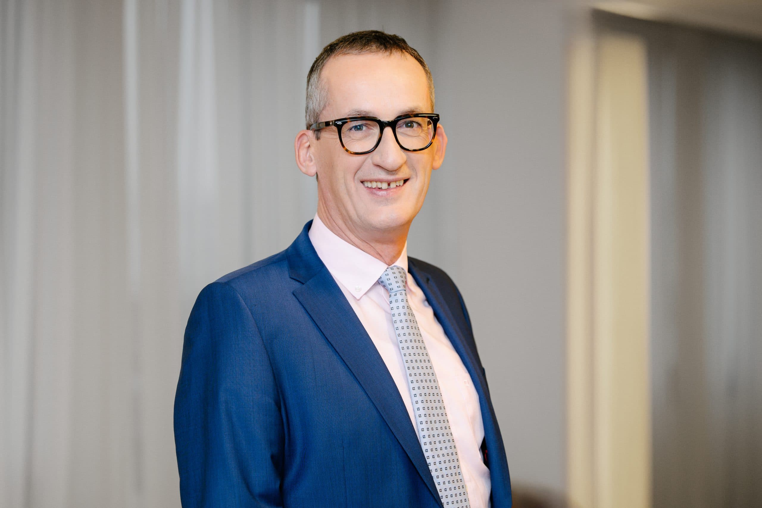 Ein Mann in einem blauen Anzug, einem hellrosa Hemd und einer gemusterten Krawatte steht in einem Haus und lächelt in die Kamera. Er hat kurze Haare, trägt eine Brille und steht vor einem sanft beleuchteten Hintergrund mit vertikalen Jalousien.