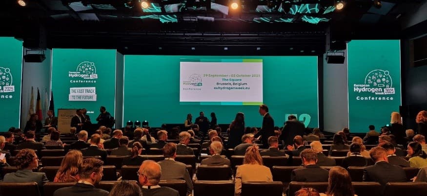 Ein großes Publikum steht in einem modernen Konferenzsaal vor einer Bühne mit türkisfarbenen Bildschirmen, auf denen 'European Hydrogen 2023 Conference' und Veranstaltungsdetails angezeigt werden. Die Menschen sitzen und stehen und bereiten sich auf den Beginn der Veranstaltung vor.