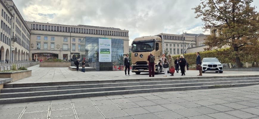 Eine Gruppe von Menschen steht in der Nähe eines goldenen Lastwagens und eines weißen Autos, die auf einem Stadtplatz mit großen Steinbauten und einem Baum im Hintergrund geparkt sind. Eine gläserne Struktur mit einem Banner ist ebenfalls zu sehen.