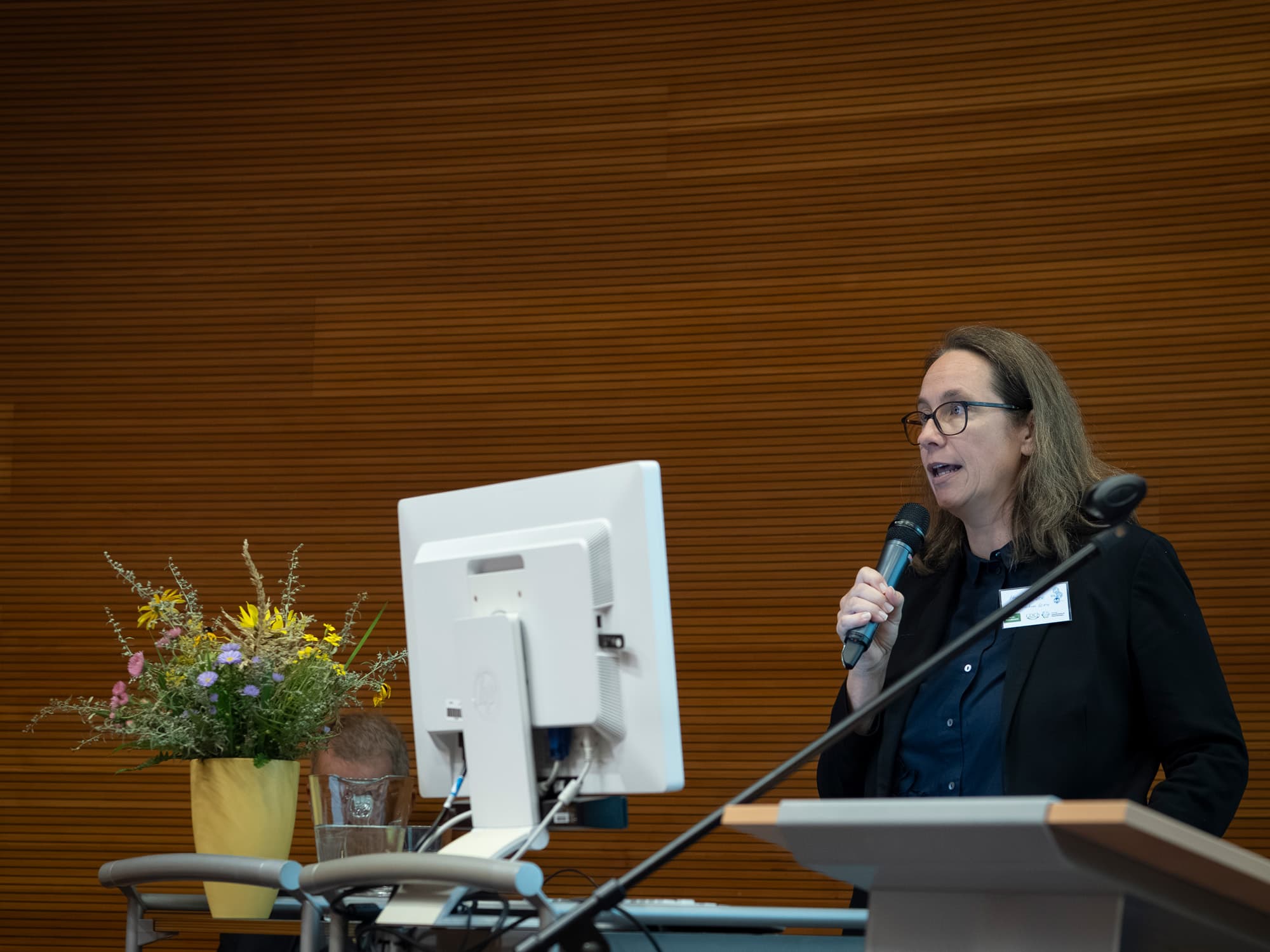 Eine Frau mit Brille und dunklem Blazer spricht an einem Podium in einem modernen Hörsaal in ein Mikrofon. Neben ihr sind ein Blumenstrauß in einer gelben Vase und ein Computermonitor zu sehen.