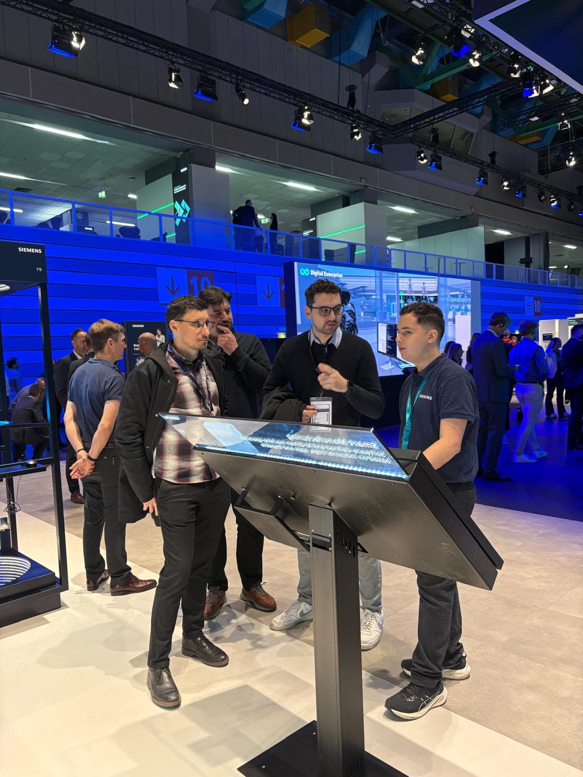 Vier Männer stehen auf einer Technologieausstellung um ein interaktives Display herum, unterhalten sich und sehen beschäftigt aus. Im hell erleuchteten Hintergrund sind andere Teilnehmer und Stände zu sehen.