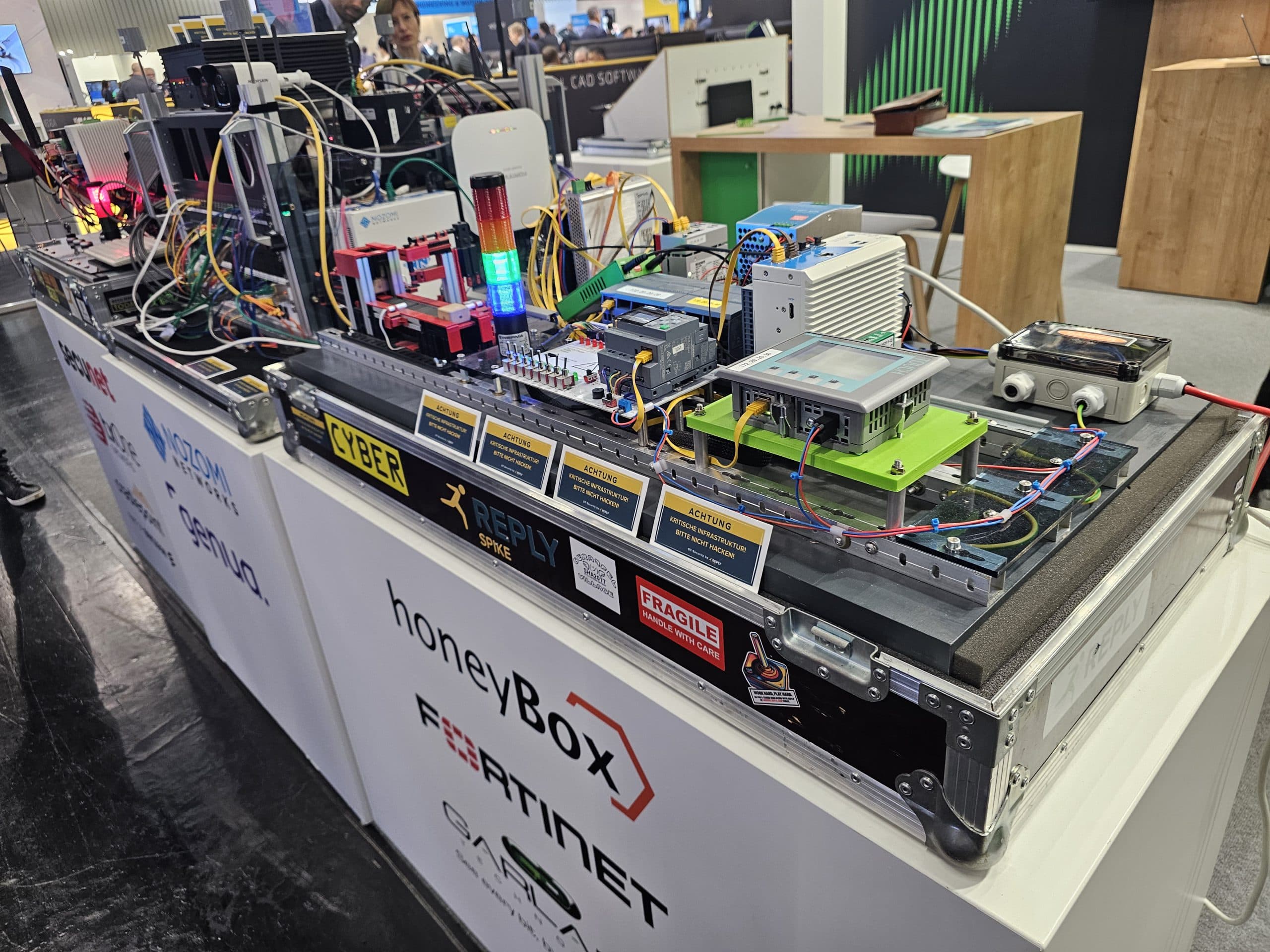 Ein Ausstellungstisch auf einer Technologiemesse zeigt elektronische Hardware, Leiterplatten, bunte Anzeigeleuchten, Bildschirme und verschiedene Etiketten, die Lösungen für Cybersicherheit und industrielle Automatisierung präsentieren.