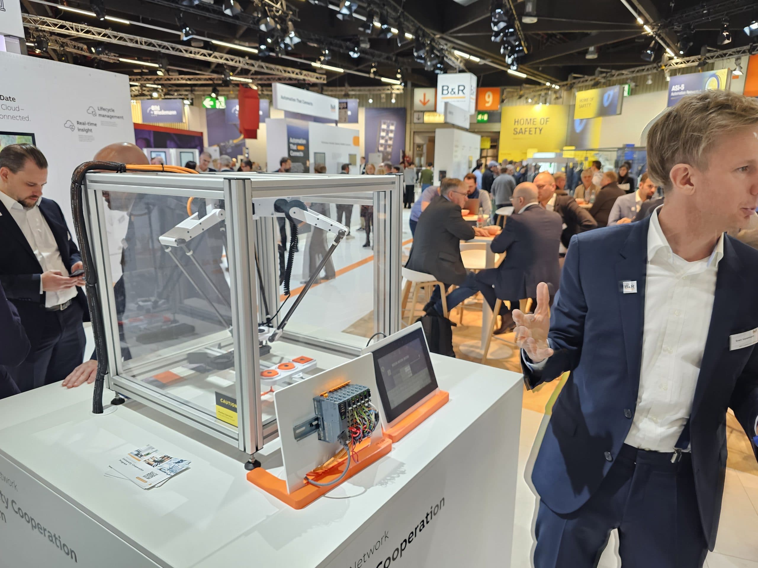Eine Geschäftsausstellungsszene mit Menschen in Anzügen, die um Stände herum Networking betreiben. Im Vordergrund steht ein Roboter in einem Glasgehäuse mit einem Bedienfeld und einem Bildschirm, der auf einem weißen Ständer steht.