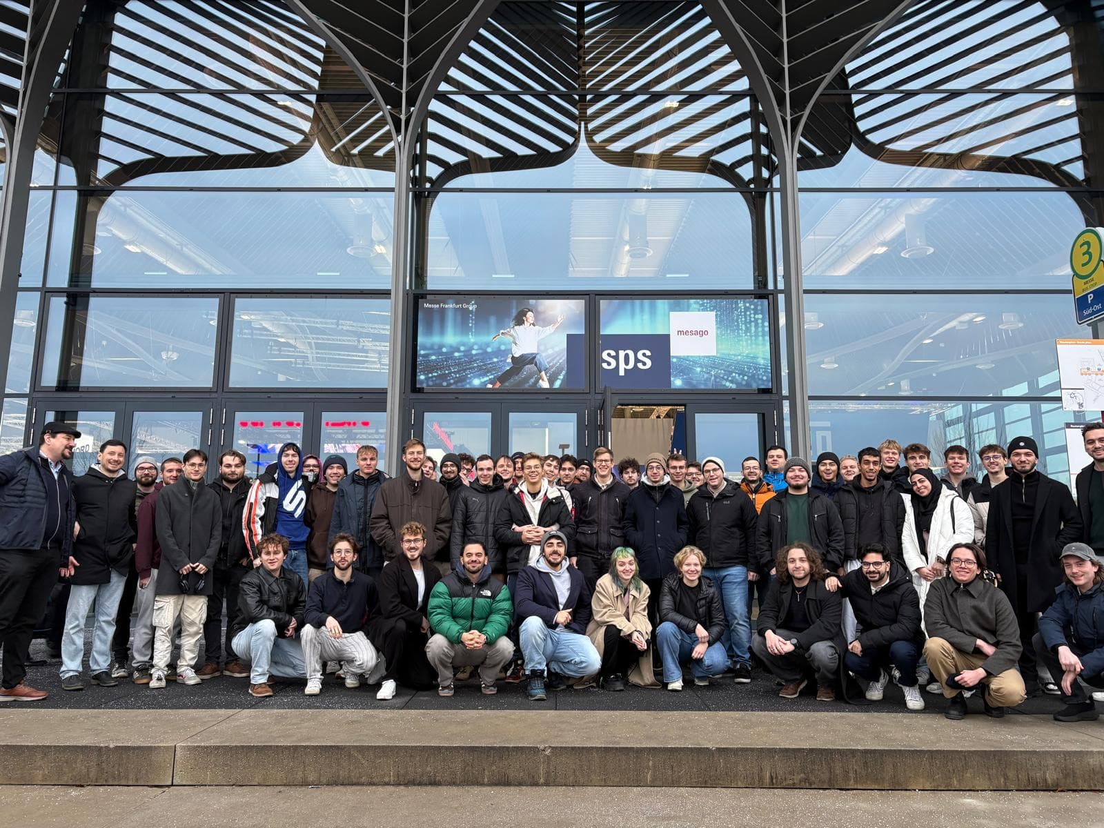 Eine große Gruppe von Menschen posiert für ein Foto vor einem modernen Glasgebäude mit den Schildern