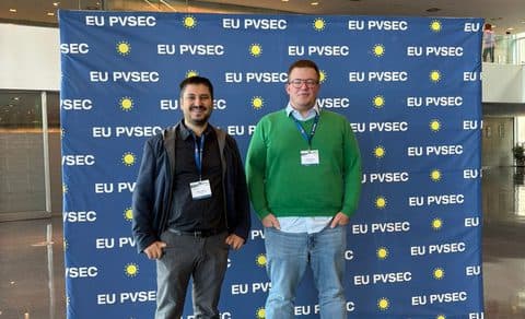 Zwei Personen mit Konferenzabzeichen stehen lächelnd vor einem blauen Stufenbanner mit gelben Sonnen und der Aufschrift EU PVSEC in einem modernen Gebäude mit Glaswänden.