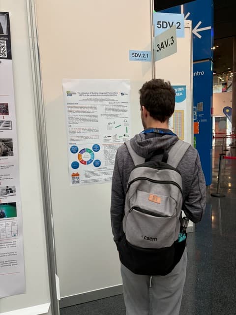 Eine Person mit kurzen lockigen Haaren, die einen grauen Kapuzenpulli und einen Rucksack trägt, steht vor einem wissenschaftlichen Poster, das bei einer Konferenz oder Ausstellung in einer Halle auf einer weißen Tafel angebracht ist.