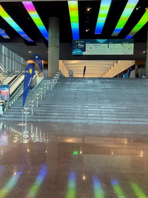 Breites Treppenhaus in einem modernen Gebäude mit regenbogenfarbenen Lichtern an der Decke, Fahnen auf beiden Seiten der Treppe und einem großen Willkommensbanner am oberen Ende. Der Boden spiegelt die Farben von oben wider.
