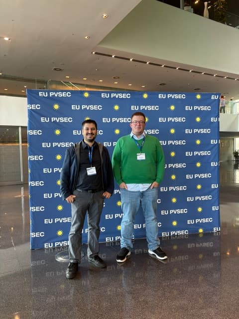 Zwei Männer stehen lächelnd vor einem blauen EU PVSEC-Hintergrund mit gelben Sonnensymbolen bei einer Indoor-Veranstaltung. Beide tragen Konferenzabzeichen und legere Kleidung. Die Umgebung wirkt modern, mit Glas und hohen Decken.