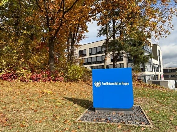 Ein blaues Schild mit dem Logo und Text FernUniversität in Hagen steht auf einer Wiese mit Herbstlaub, vor einem modernen Gebäude und Bäumen mit Herbstlaub.