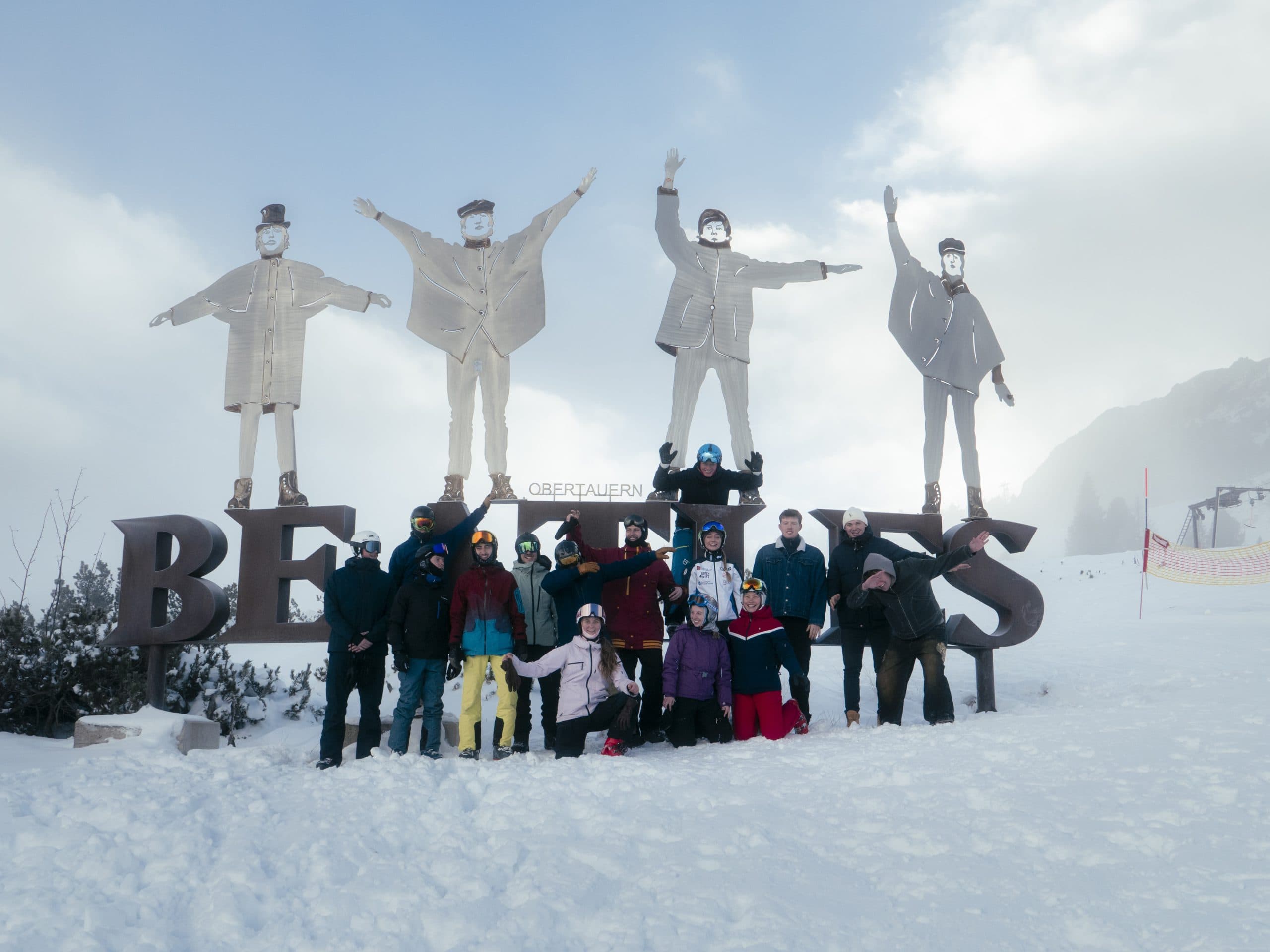 Eine Gruppe von Menschen in Winterkleidung posiert vor großen Metallstatuen und dem Wort "Beatles" im Schnee, mit einem bewölkten Himmel und Bergen im Hintergrund.