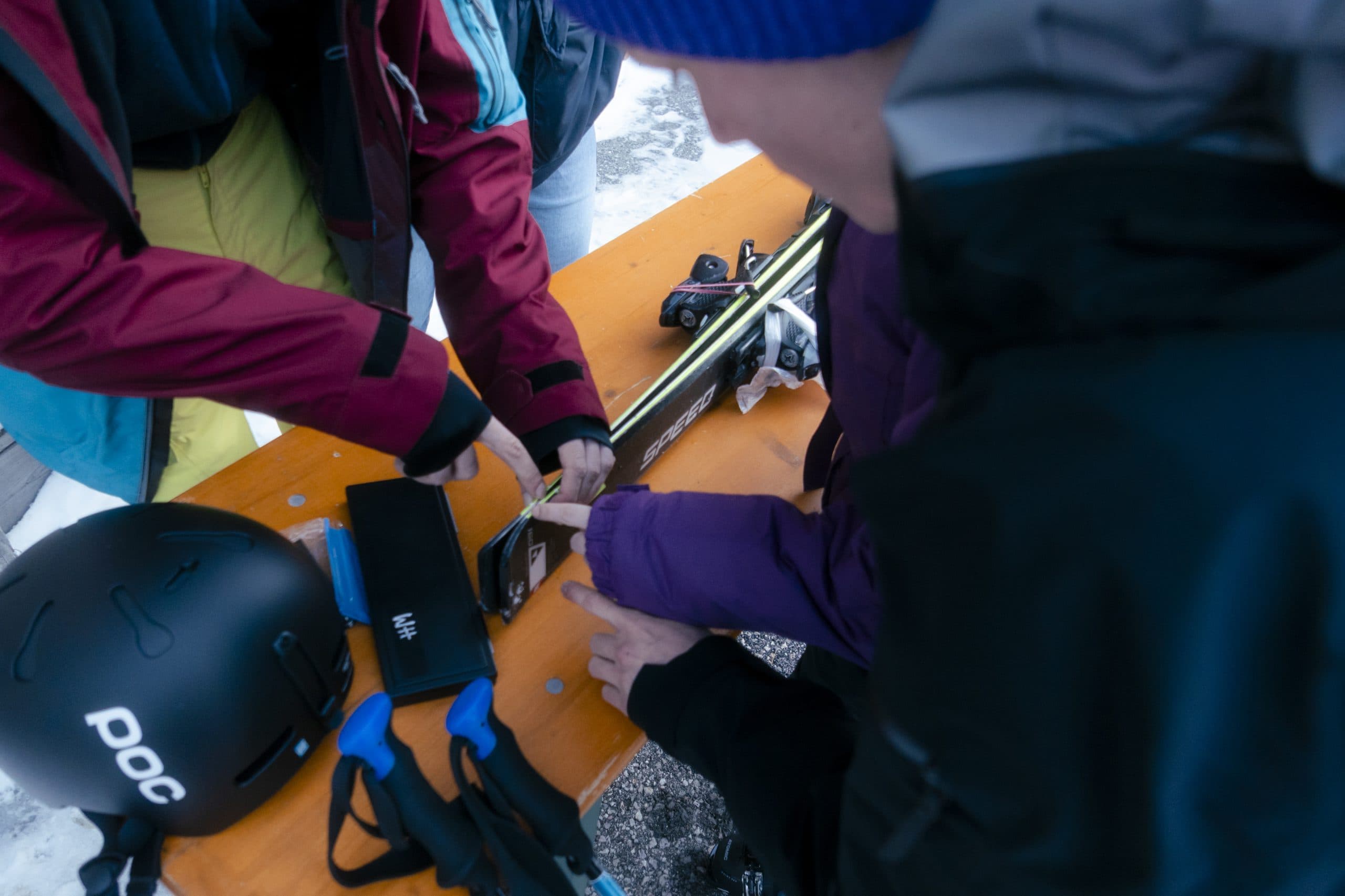 Drei Personen sind um einen Holztisch versammelt und stellen eine Skibindung ein. Ein schwarzer Helm und Skistöcke liegen auf dem Tisch, und die Szene scheint im Freien in einer verschneiten Gegend zu spielen.