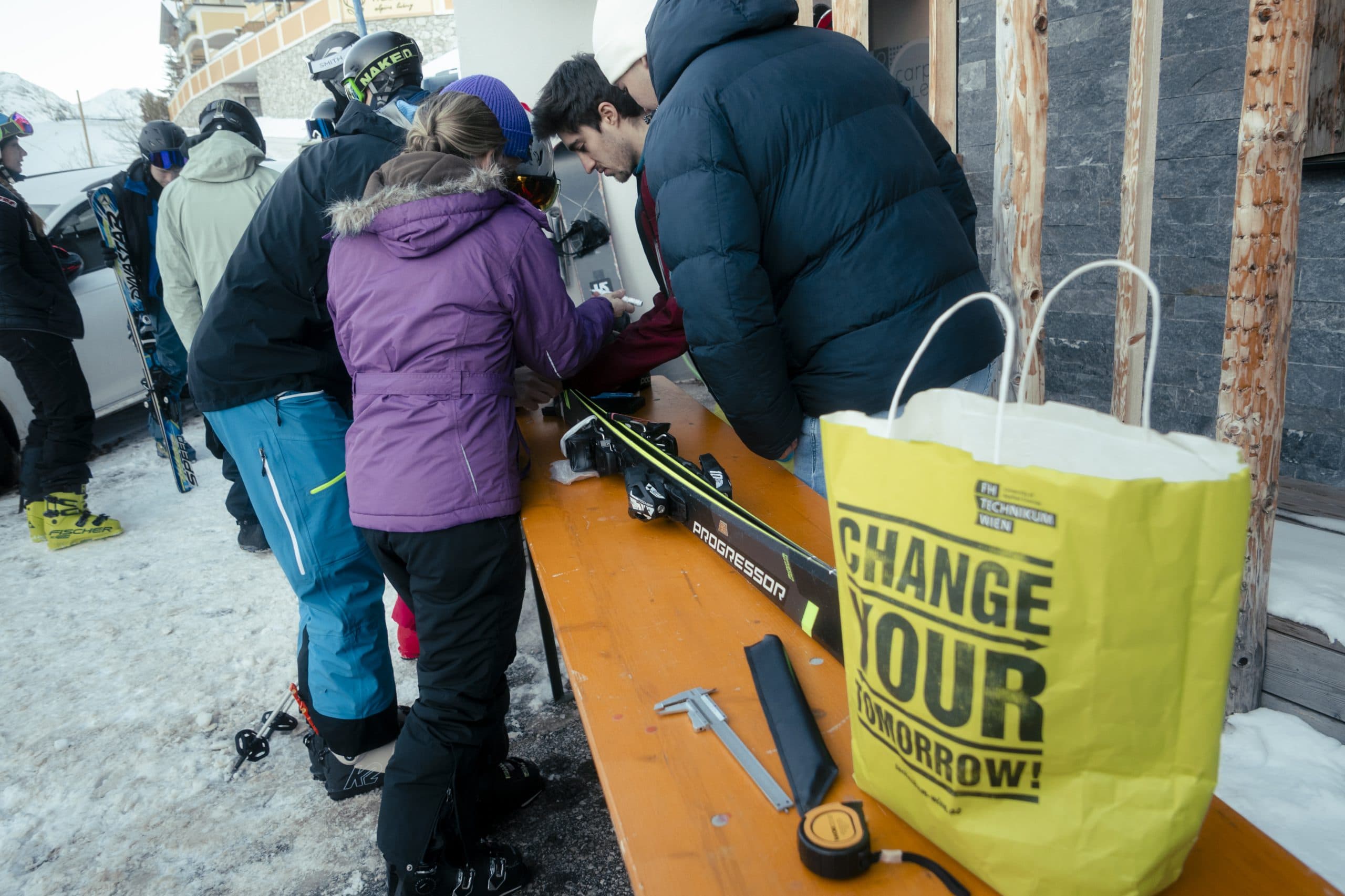 Eine Gruppe von Menschen in Winterkleidung versammelt sich draußen im Schnee um einen Holztisch mit einer Skiausrüstung. Auf dem Tisch liegen eine gelbe Tasche mit der Aufschrift CHANGE YOUR TOMORROW! und Werkzeug. Im Hintergrund sind Skifahrer und Gebäude zu sehen.