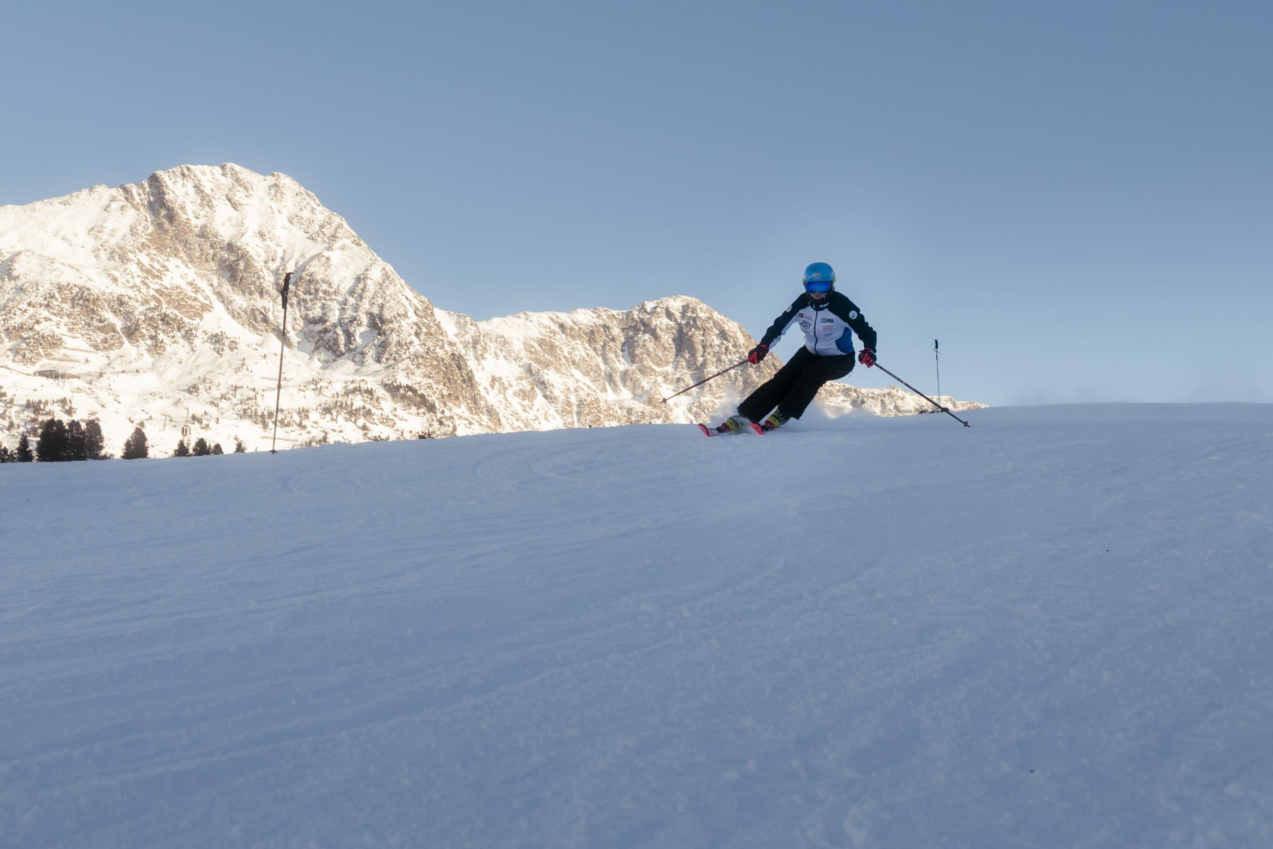 Ein Skifahrer mit blauem Helm und blauer Jacke fährt auf einer verschneiten Piste mit schneebedeckten Bergen im Hintergrund unter einem klaren blauen Himmel bergab.