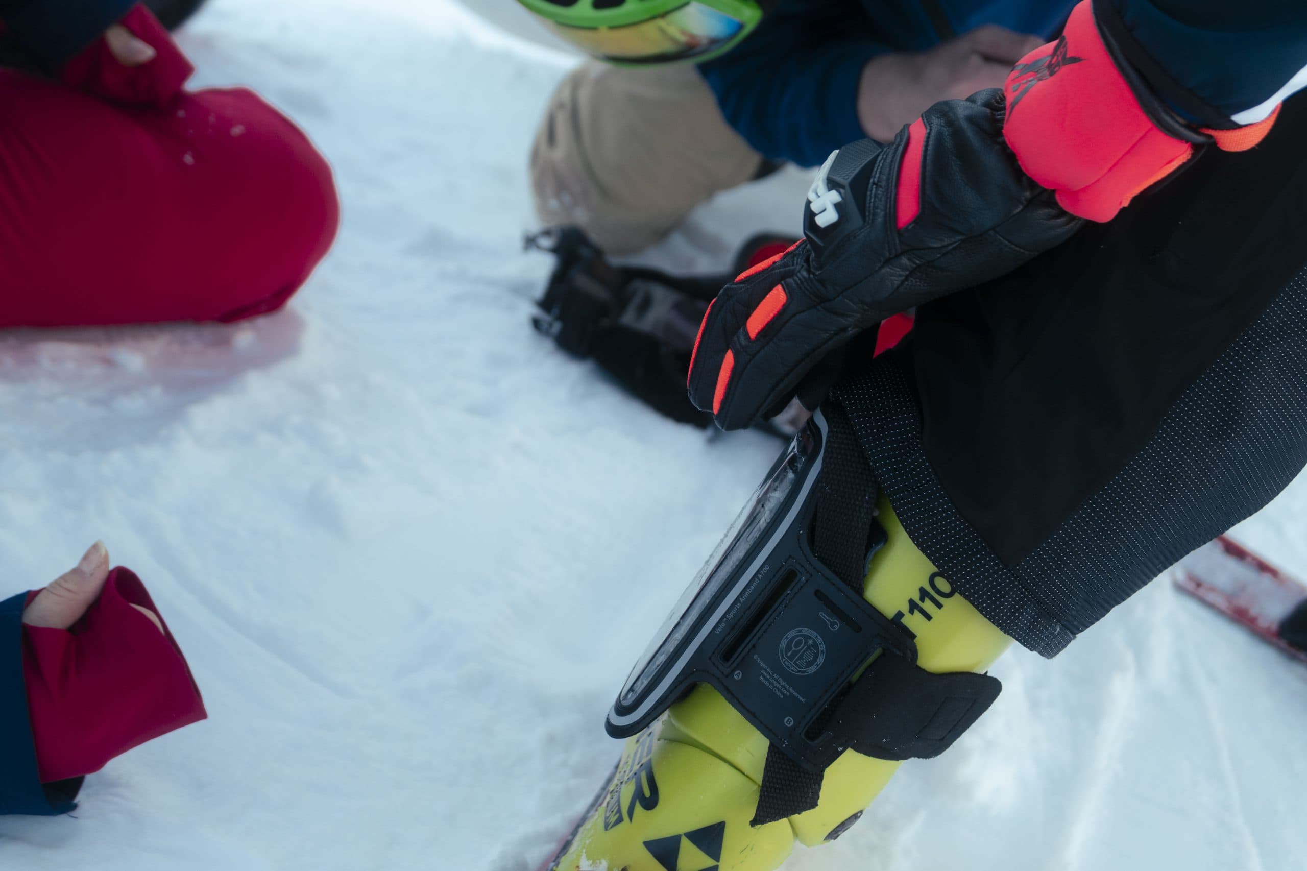 Nahaufnahme eines Skifahrers mit Handschuhen, Skiausrüstung und einem am Bein befestigten Funkgerät, der im Schnee kniet, während eine andere Person in roten Hosen ebenfalls in der Nähe kniet.