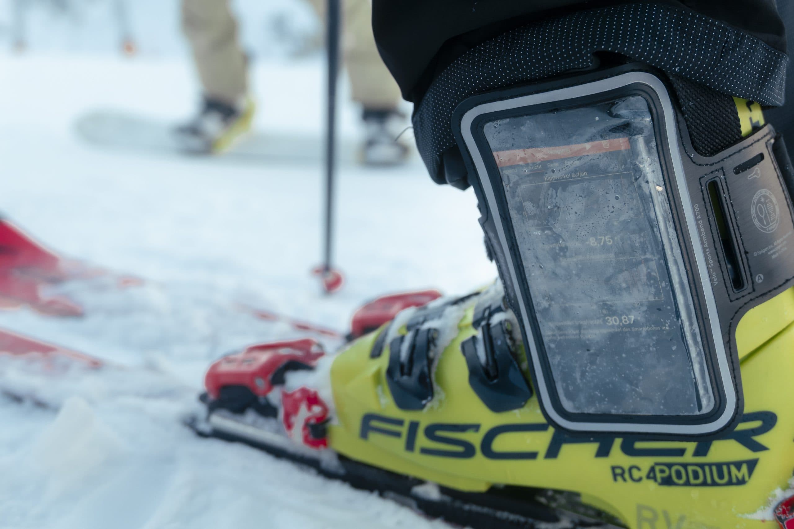 Nahaufnahme des gelben Fischer-Skischuhs eines Skifahrers mit einem am Knöchel befestigten Gerät, das Zahlen anzeigt. Der Boden ist mit Schnee bedeckt, und im Hintergrund ist eine weitere Person mit Skiern und Stöcken zu erkennen.
