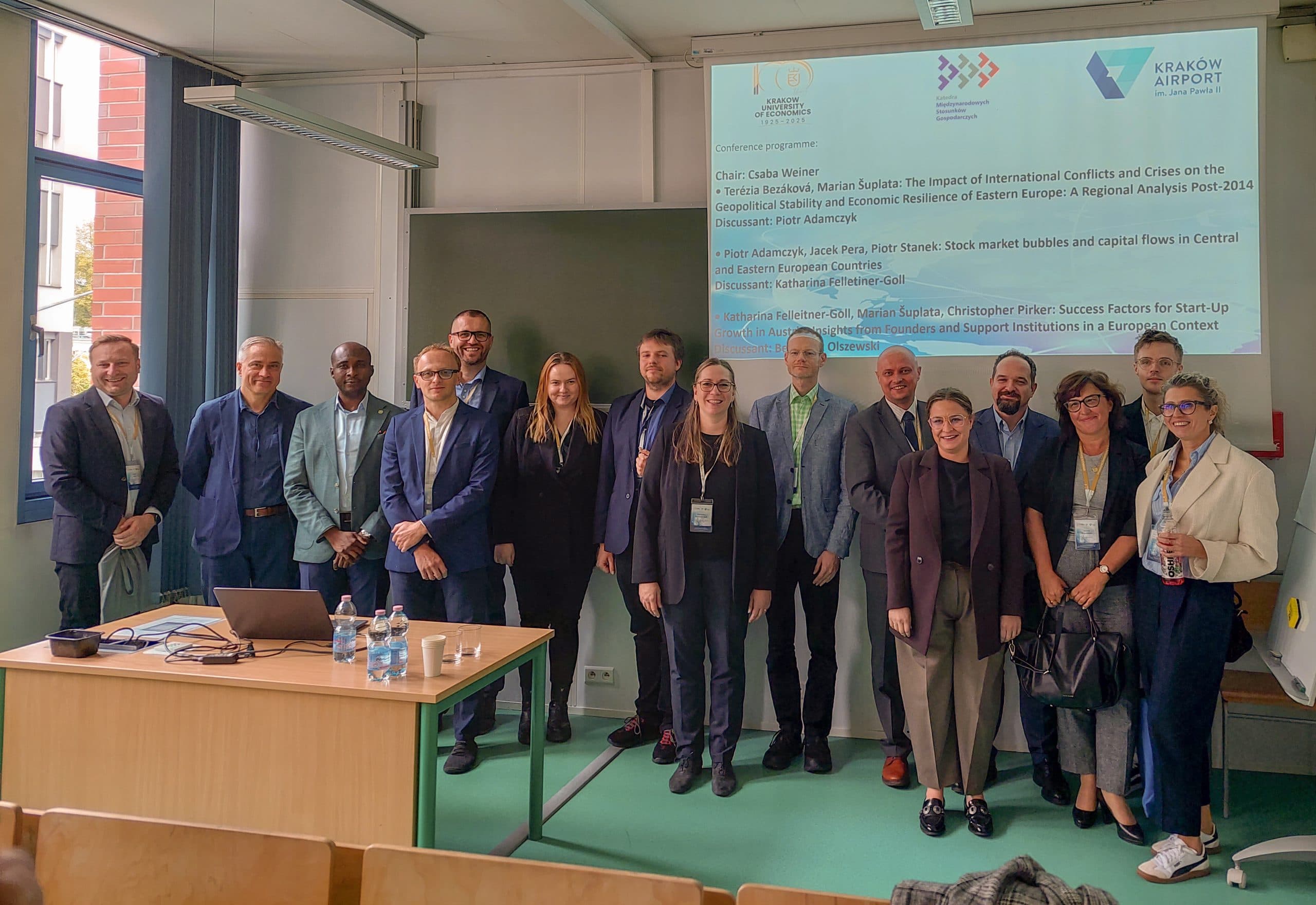 Eine Gruppe von fünfzehn Personen, gekleidet in Business- oder eleganter Freizeitkleidung, steht gemeinsam vor einem Klassenzimmer. Hinter ihnen wird eine Präsentationsfolie an die Wand projiziert, und vor ihnen steht ein Tisch mit Wasserflaschen.