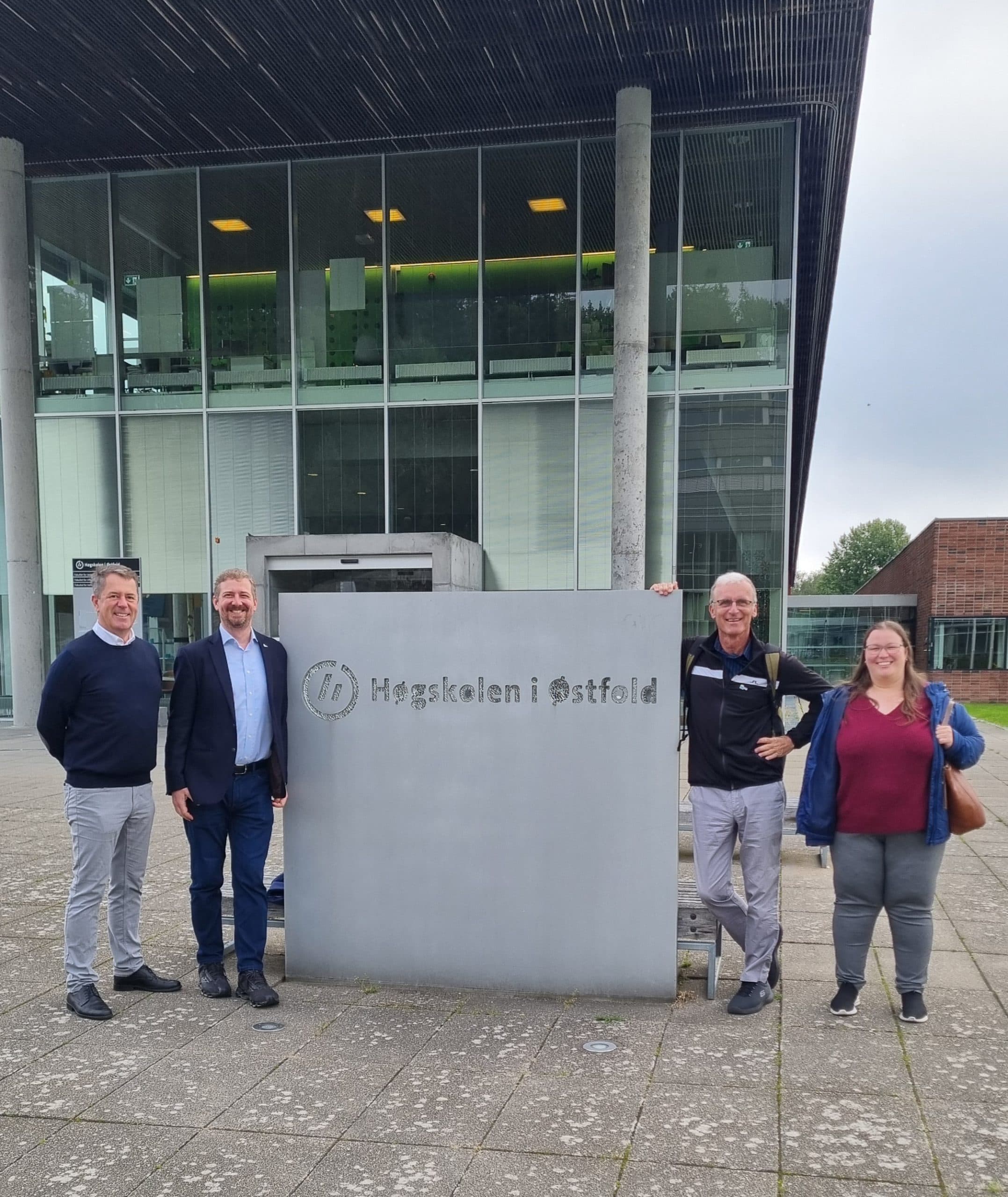 Vier Personen stehen draußen vor einem Schild mit der Aufschrift Høgskolen i Østfold. Das moderne Gebäude hinter ihnen hat große Glasfenster und hohe Säulen. Die Gruppe lächelt und posiert für das Foto.