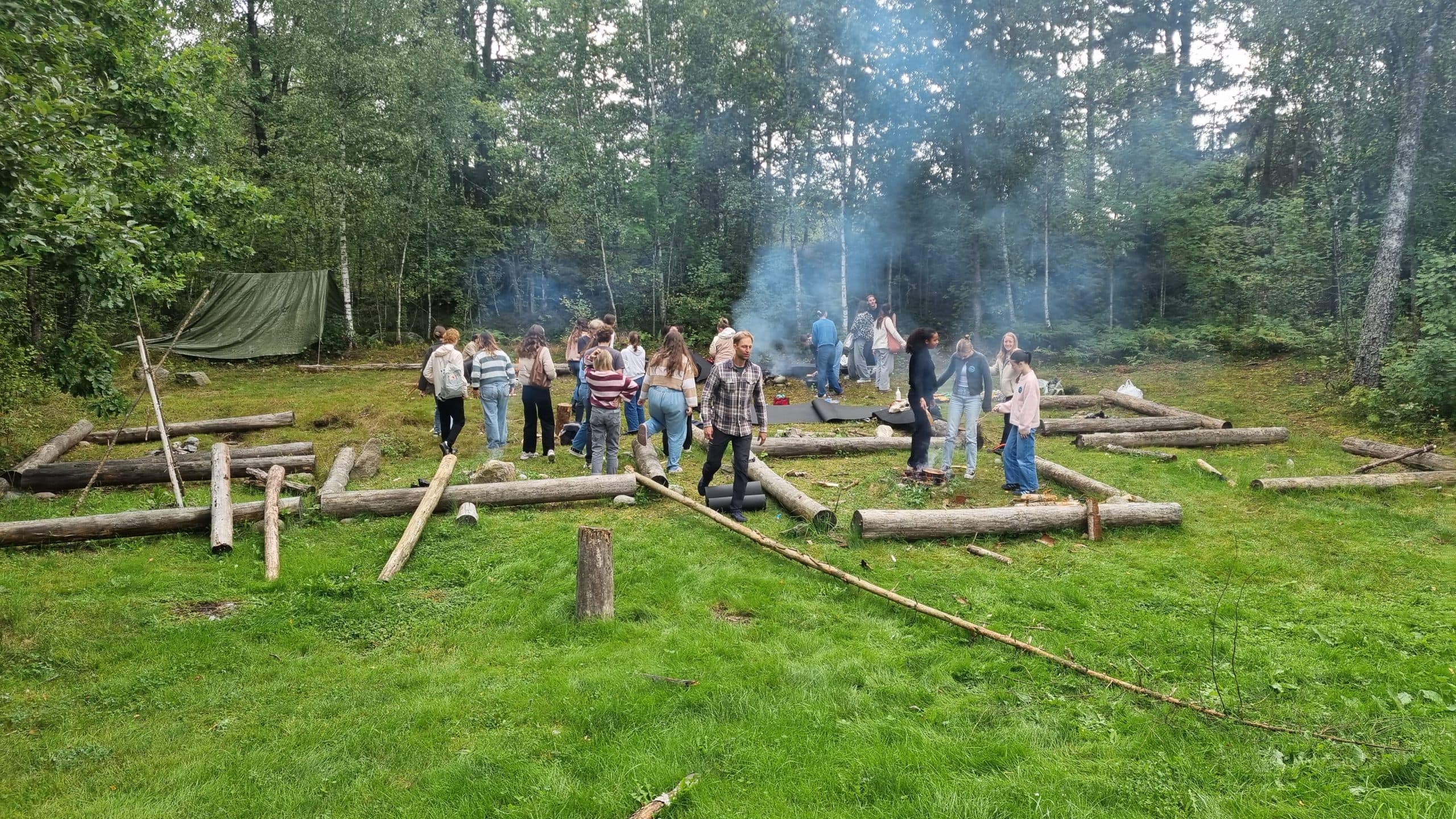 Eine Gruppe von Menschen steht und geht zwischen verstreuten Baumstämmen und Rauch auf einer grasbewachsenen, von Wald umgebenen Lichtung; einige interagieren, während andere beobachten, mit Bäumen und einer grünen Plane im Hintergrund.
