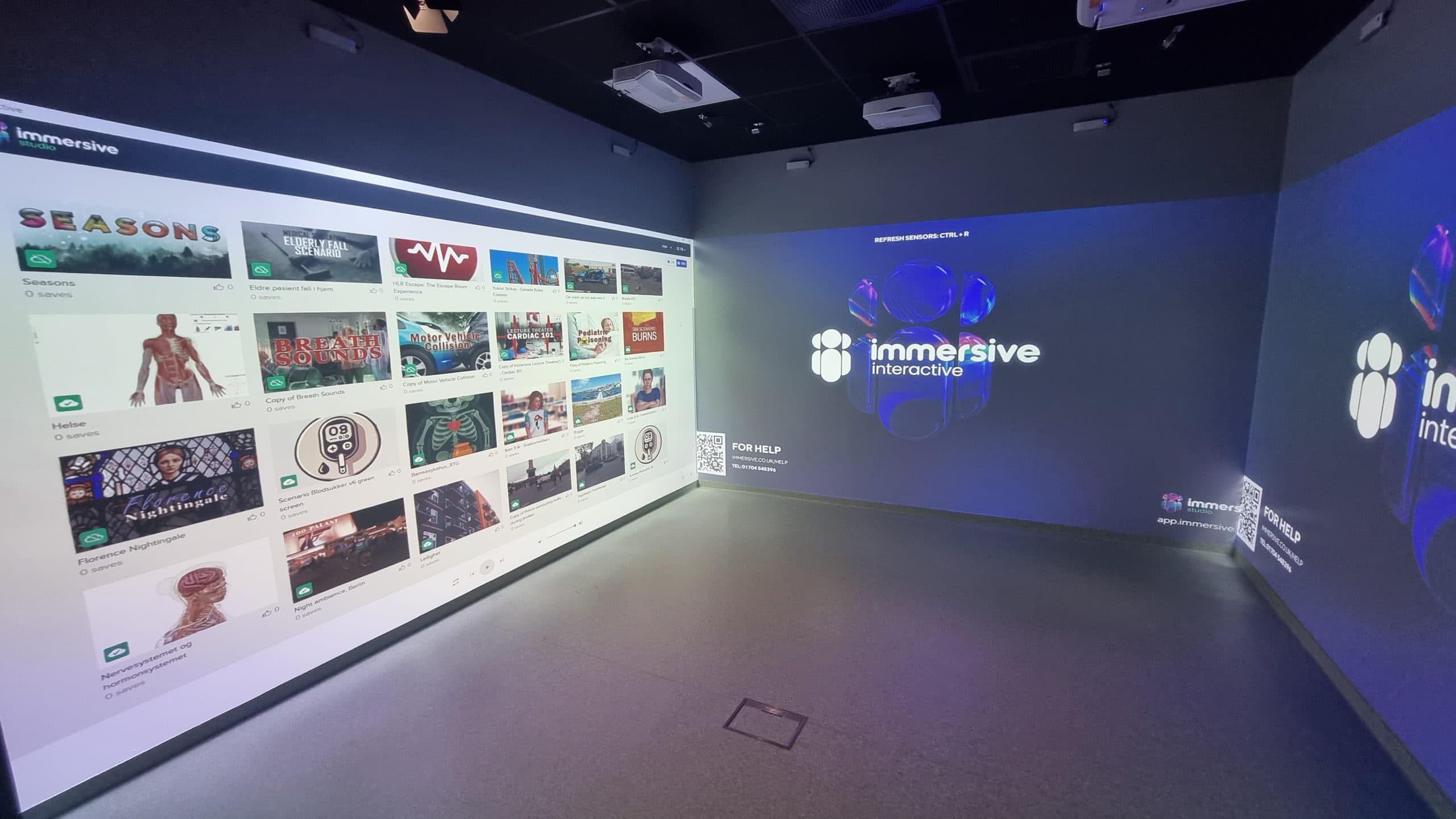 Ein Raum mit drei Projektionswänden, die eine interaktive Schnittstelle für Immersive Interactive zeigen, mit verschiedenen digitalen Inhaltsoptionen und einem leuchtenden Logo auf der zentralen Wand.