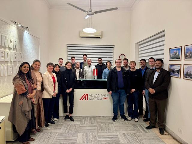 Eine Gruppe von 18 Personen steht lächelnd in einer Bürohalle in der Nähe eines Empfangsschalters mit einem Advantage Austria-Schild. Einige tragen Geschäftskleidung, andere sind leger gekleidet. An der Wand hängen mehrere gerahmte Fotos.