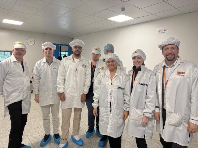 Eine Gruppe von acht Personen in weißen Laborkitteln, Haarnetzen und blauen Schuhüberzügen steht in einem sauberen, hellen Raum mit einer Uhr an der Wand zusammen. Einige Kittel sind mit VISITOR und ANANTY beschriftet. Sie lächeln in die Kamera.