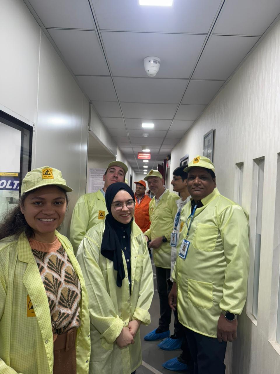 Eine Gruppe von Personen mit gelben Laborkitteln und Ausweisen steht in einem schmalen Flur, einige lächeln in die Kamera. Der Flur hat weiße Wände und Deckenplatten.