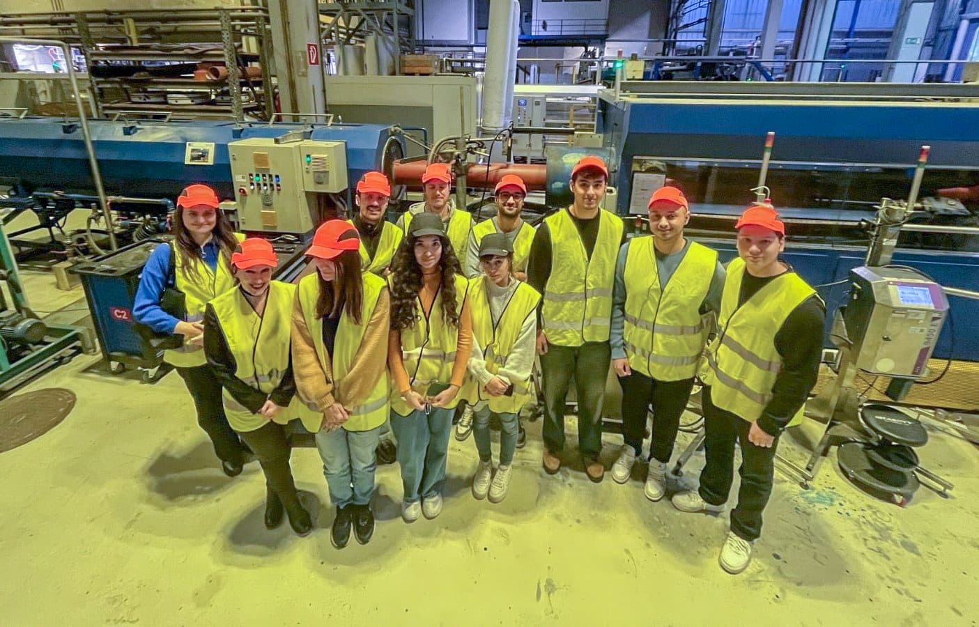 Eine Gruppe von 13 Personen, die gelbe Sicherheitswesten und rote Mützen tragen, steht in einer Fabrik- oder Industrieumgebung und posiert und lächelt für die Kamera vor großen Maschinen und Geräten.