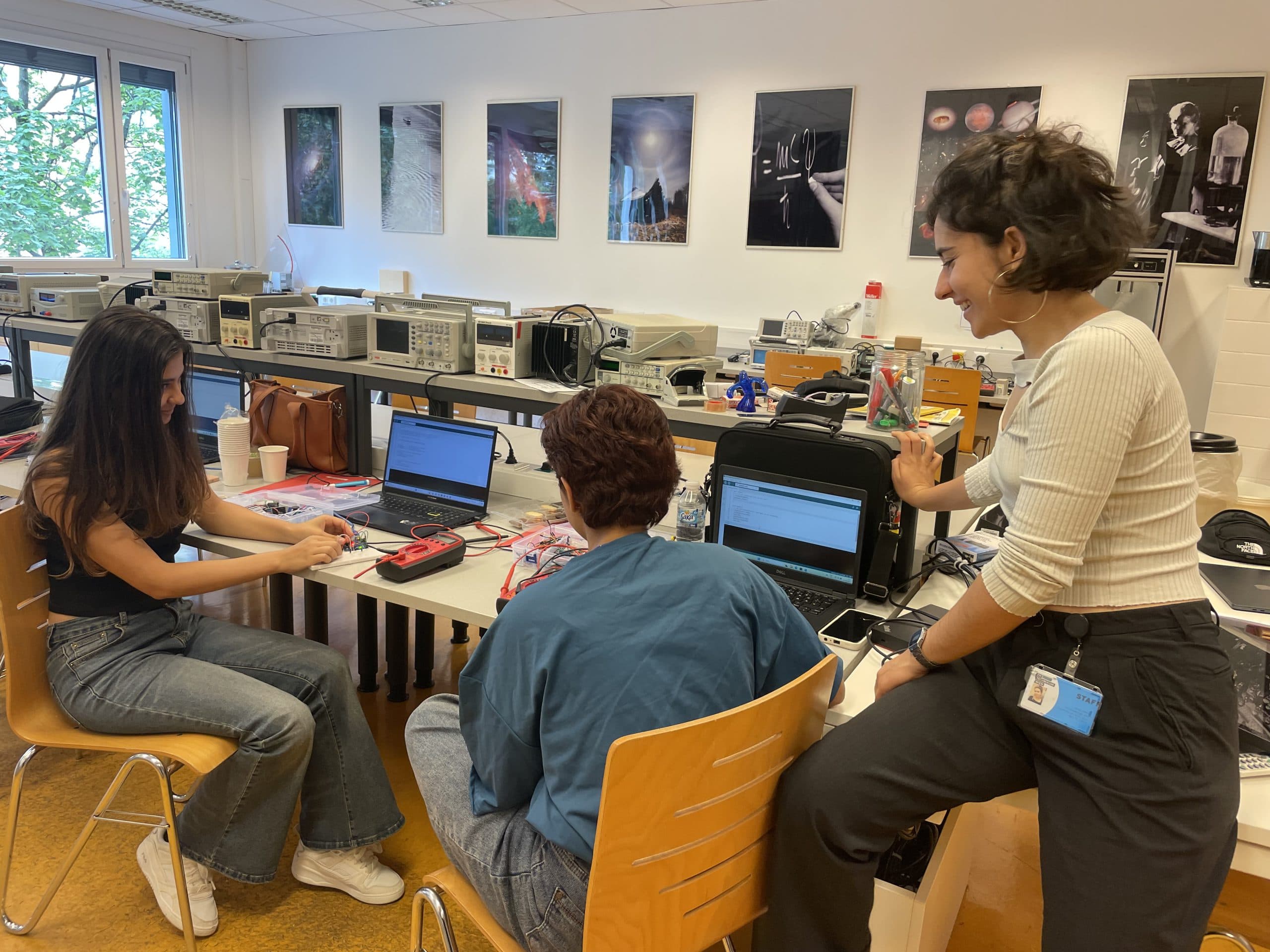 Drei Personen arbeiten zusammen an einem Tisch mit Laptops und elektronischen Geräten in einem hellen Klassenzimmer oder Labor, lächeln und diskutieren über ihr Projekt. Hinter ihnen hängen wissenschaftliche Poster an den Wänden.