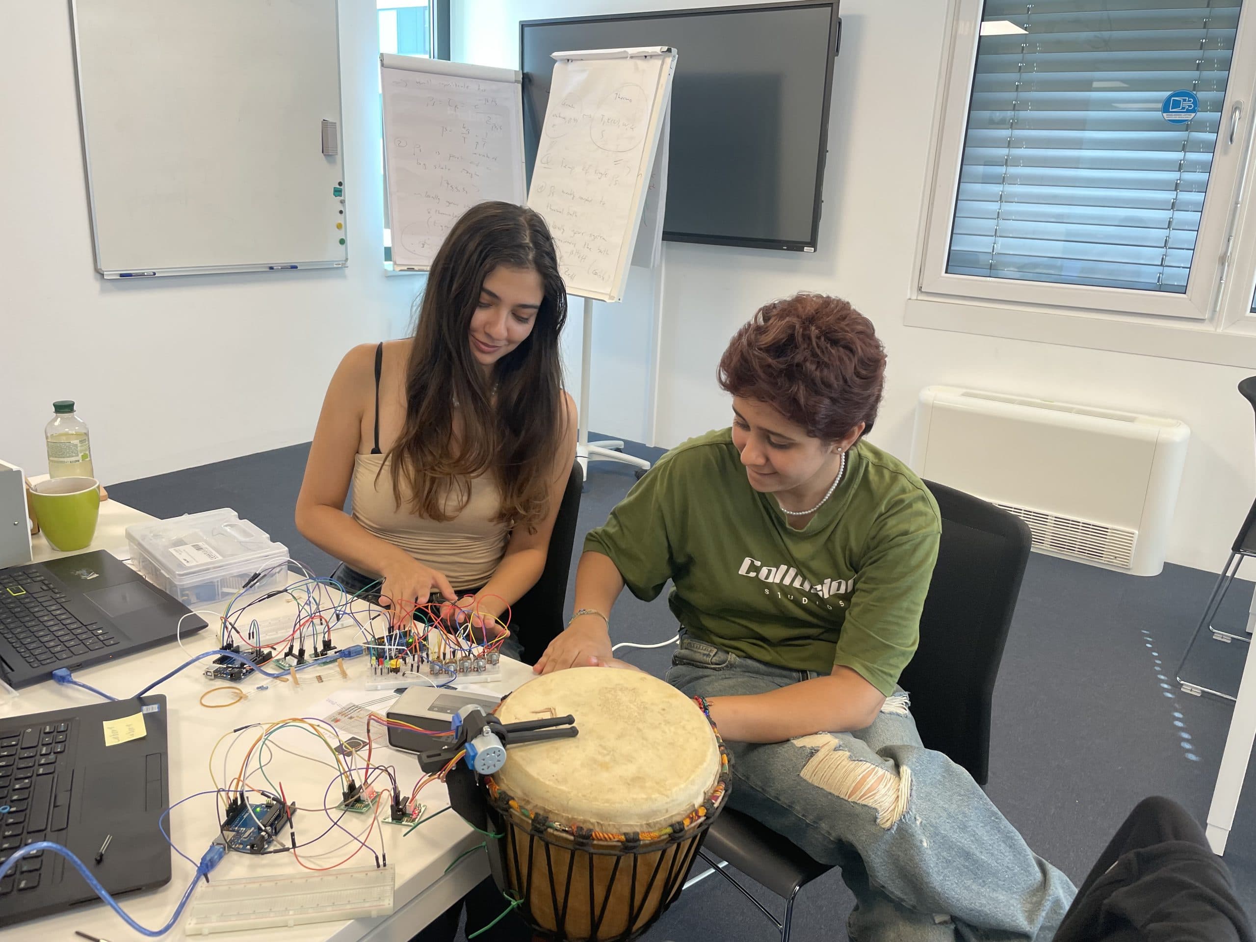 Zwei Personen sitzen an einem Tisch mit elektronischen Bauteilen und Kabeln. Eine Person klopft auf eine Trommel, während die andere mit der Elektronik interagiert. Sie scheinen konzentriert und engagiert an einem gemeinsamen Projekt zu arbeiten.