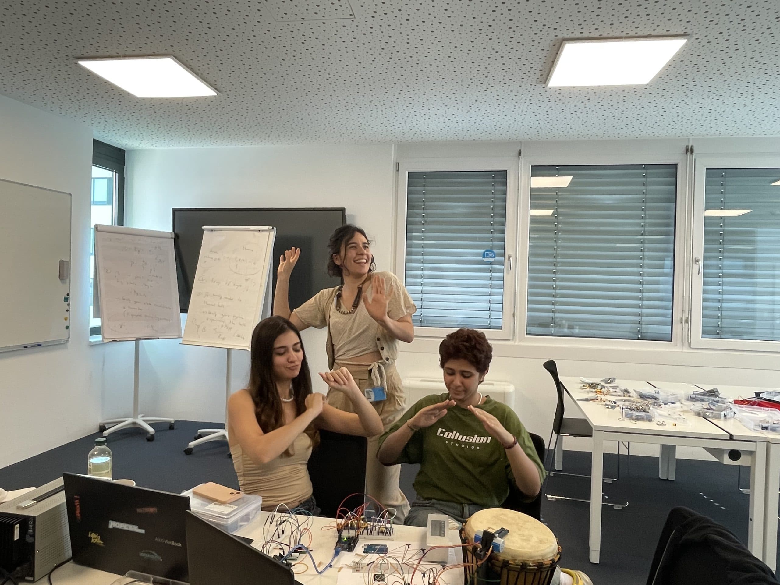 Drei junge Leute in einem modernen Klassenzimmer, zwei sitzen an einem Tisch mit Elektronik und einer Trommel, einer steht hinter ihnen, alle lächeln und tanzen. Im Hintergrund sind Whiteboards mit Notizen zu sehen.