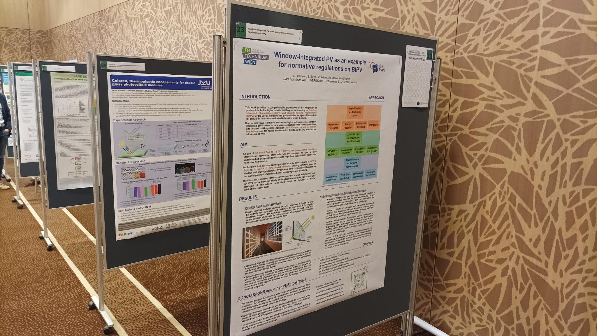 Eine Reihe von wissenschaftlichen Postern auf grauen Tafeln in einem Konferenzraum mit gemusterten Wänden und Teppichboden. Das nächstgelegene Poster befasst sich mit fensterintegrierten PV-Anlagen im Hinblick auf normative Vorschriften für BIPV.