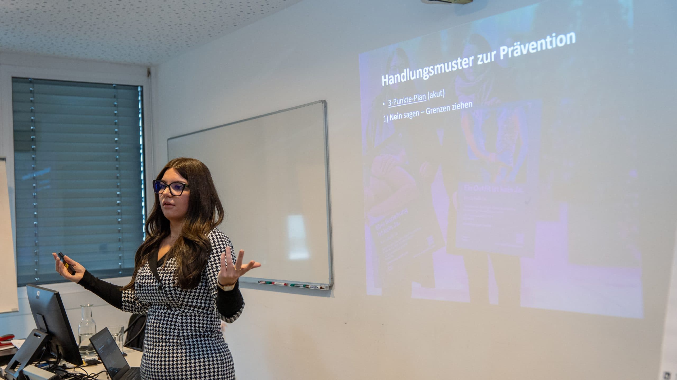 Eine Frau steht vor einem Whiteboard und einer projizierten Präsentationsfolie und gestikuliert, während sie zu einem Publikum in einem Klassenzimmer spricht. Die Folie ist auf Deutsch und trägt den Titel