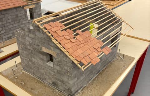 Ein maßstabsgetreues Modell eines grauen Backsteinhauses mit einem teilweise mit Ziegeln gedeckten Schrägdach, hölzernen Dachbalken und einigen im Hintergrund sichtbaren Bauwerkzeugen, das auf einem Tisch mit roten Beinen in einem Innenraum ausgestellt ist.