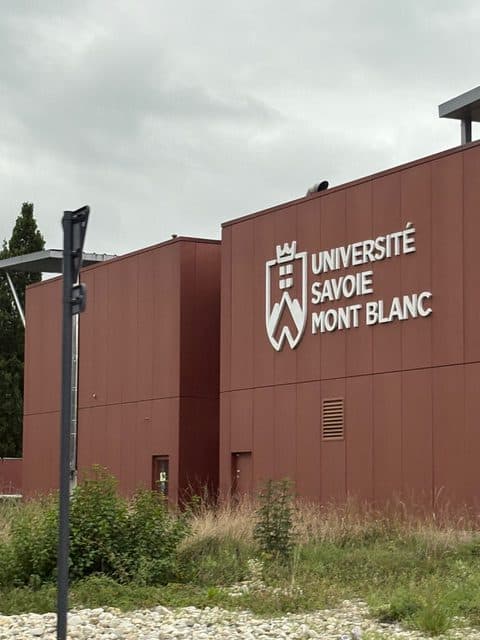 Ein rotes Gebäude mit dem Schriftzug Université Savoie Mont Blanc und seinem Logo an der Wand, umgeben von Gras und Kieselsteinen, unter einem bewölkten Himmel.