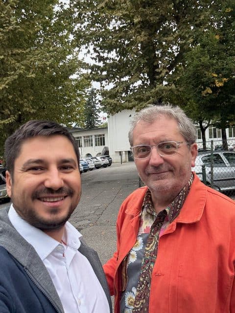 Zwei Männer lächeln im Freien vor einer Reihe von geparkten Autos und Bäumen. Ein Mann trägt ein weißes Hemd und eine graue Jacke, der andere eine Brille, eine orangefarbene Jacke und ein gemustertes Hemd. Es ist ein bewölkter Tag.