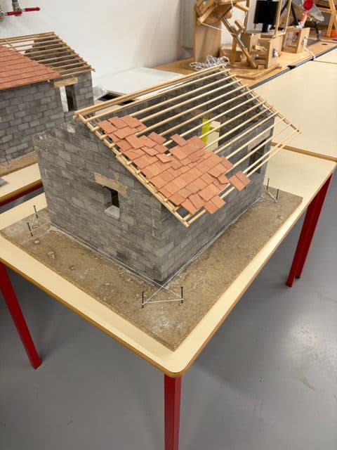 Ein Modellhaus mit grauen Ziegelwänden steht auf einem Tisch. Der hölzerne Dachstuhl ist teilweise mit roten Ziegeln bedeckt, was auf ein unfertiges Dach hinweist. In der Nähe sind im Hintergrund ein weiteres ähnliches Modell und Werkzeuge zu sehen.