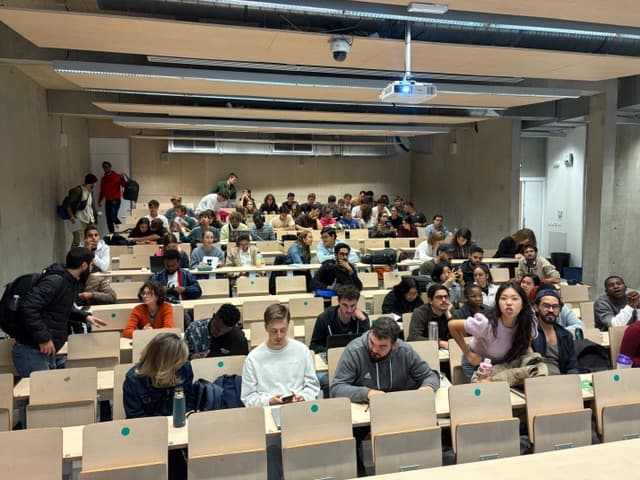 Eine große Gruppe von Studenten sitzt in einem modernen Hörsaal, unterhält sich und wartet auf den Beginn des Unterrichts. Der Raum hat Holztische, Betonwände und einen an der Decke montierten Projektor. Einige Studenten stehen oder gehen zwischen den Reihen umher.