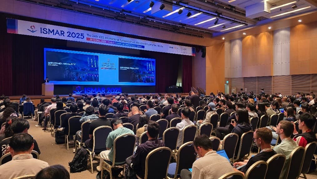 Auf der ISMAR-Konferenz 2025 sitzt ein großes Publikum in Reihen vor einer Bühne, auf der zwei große Bildschirme MIXED REALITY zeigen und ein Redner auf einem Podium spricht.