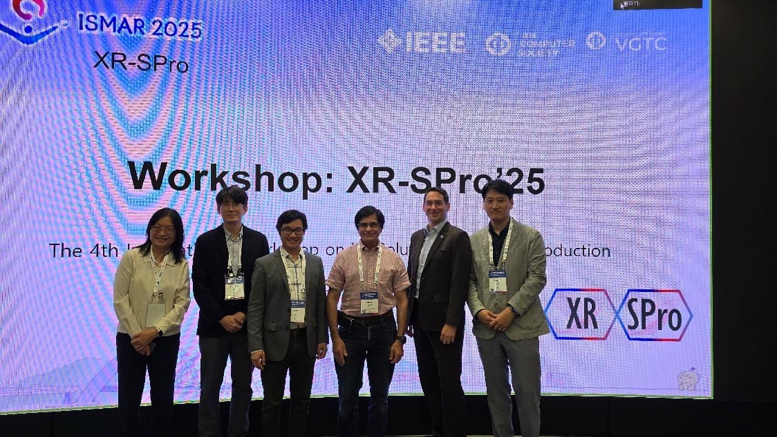 Sechs Personen stehen zusammen auf einer Bühne vor einem großen Bildschirm mit der Aufschrift 'Workshop: XR-SPro’25' auf der Veranstaltung ISMAR 2025, mit verschiedenen Logos, darunter IEEE, XR und SPro im Hintergrund.