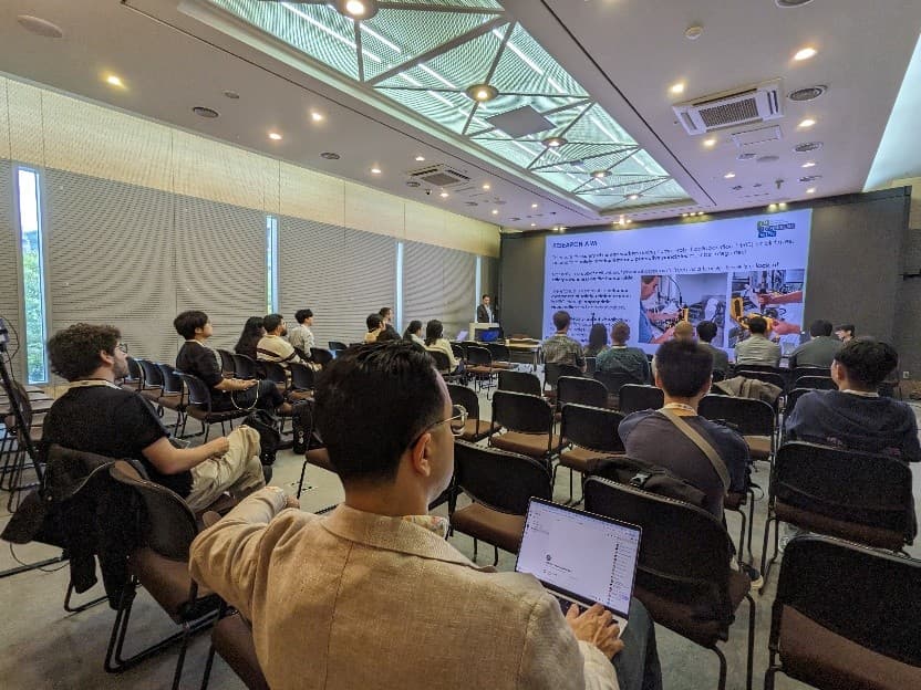 Die Teilnehmer sitzen in einem Konferenzraum und sehen zu, wie ein Redner an der Stirnseite Folien präsentiert. Der Raum hat große Fenster, Deckenbeleuchtung, und die Teilnehmer arbeiten mit Laptops und machen sich Notizen.