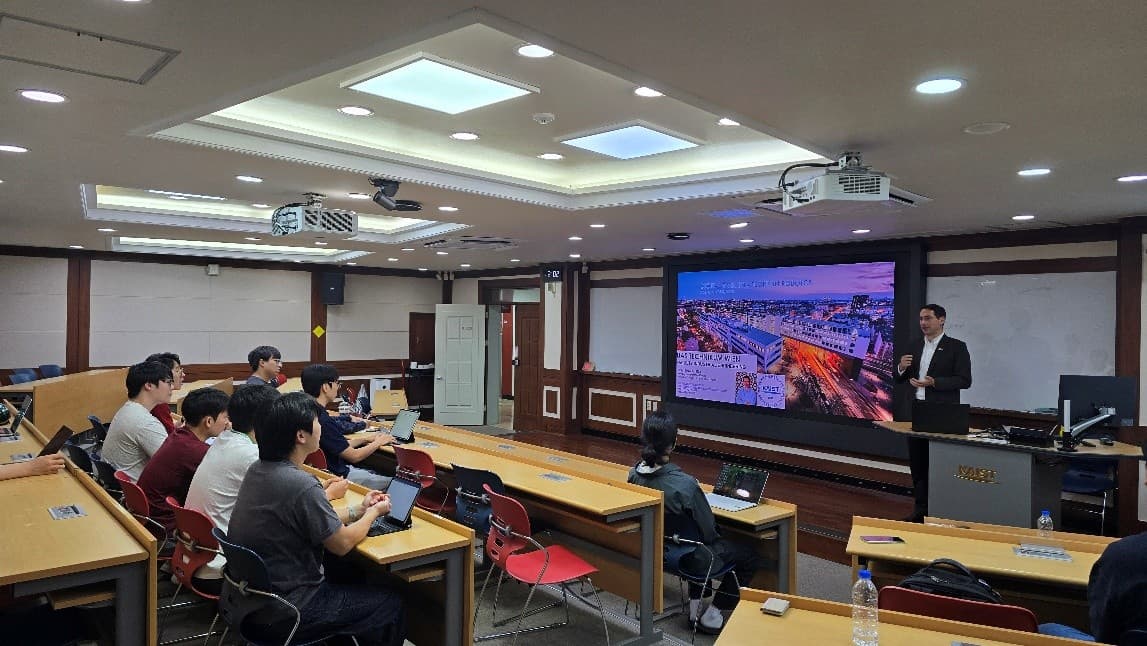 Ein Mann im Anzug hält einen Vortrag vor einem Klassenzimmer mit Schülern, die an Tischen sitzen, während hinter ihm ein großer Bildschirm eine bunte Stadtlandschaft und Text zeigt. Der Raum ist mit moderner Beleuchtung und AV-Ausrüstung ausgestattet.