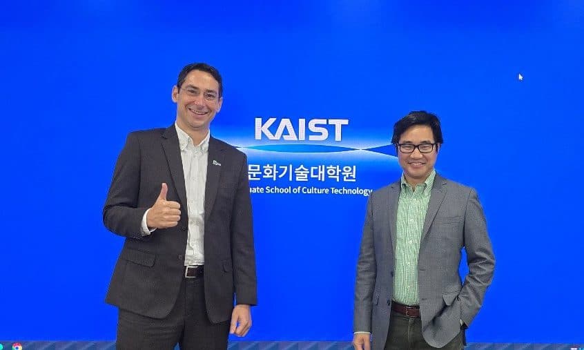 Zwei Männer in Geschäftskleidung stehen lächelnd vor einem großen Bildschirm, auf dem das Logo und der Text des KAIST zu sehen sind. Einer der Männer zeigt den Daumen nach oben. Die Wände des Raums sind mit Holz verkleidet.