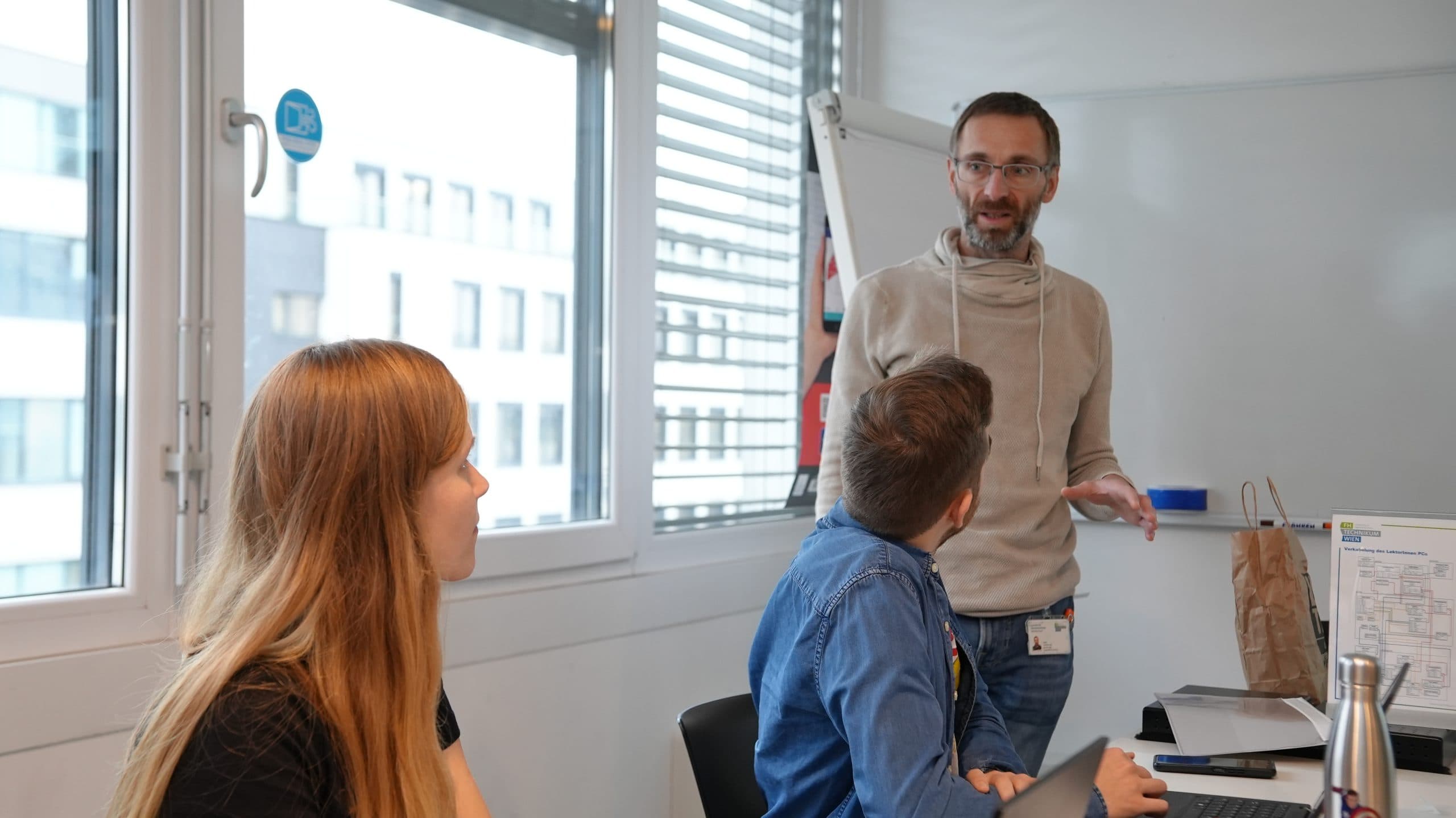 Drei Personen führen eine Diskussion in einem modernen Büro. Ein Mann steht und spricht, während zwei andere, eine Frau und ein weiterer Mann, an einem Tisch mit Laptops sitzen und aufmerksam zuhören. Große Fenster sorgen für natürliches Licht.
