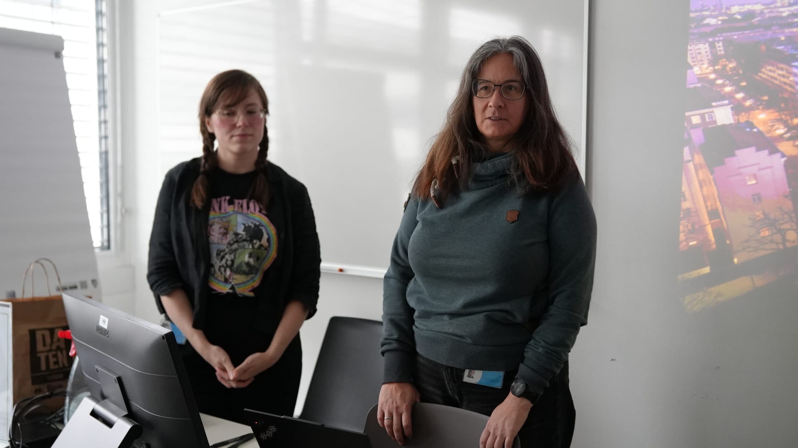 Zwei Frauen stehen in einem Klassenzimmer oder Büro, die eine an einem Laptop, die andere neben ihr. An der Wand ist eine Projektion einer Stadtlandschaft zu sehen und im Hintergrund ein Whiteboard.